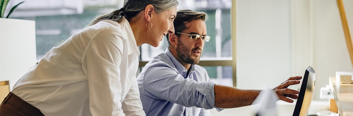 Two colleagues collaborating at a desk in a modern office, with one person pointing at a computer monitor while the other leans in to observe.