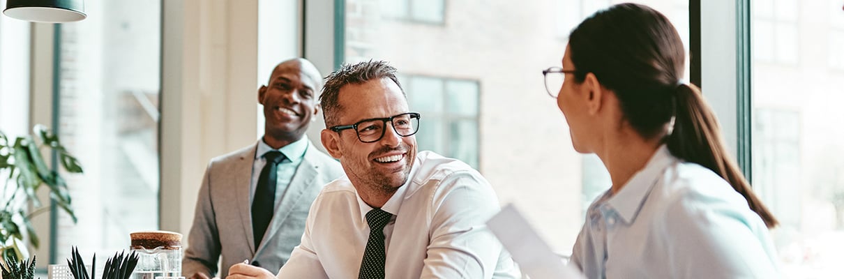 Three professionals in a modern office setting engaged in a discussion around a table, with documents and plants visible in the foreground