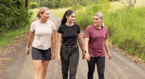 Three women walking together on a nature trail surrounded by green trees and vegetation