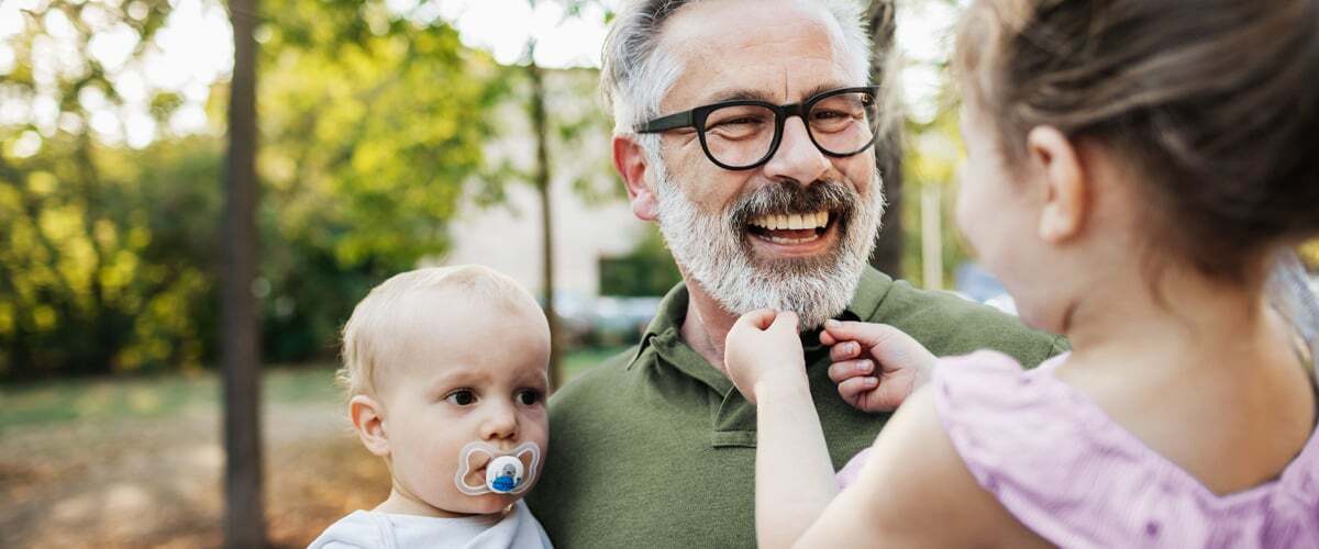 An older man with a white beard carrying a small child. A young girl is grabbing the man's beard.