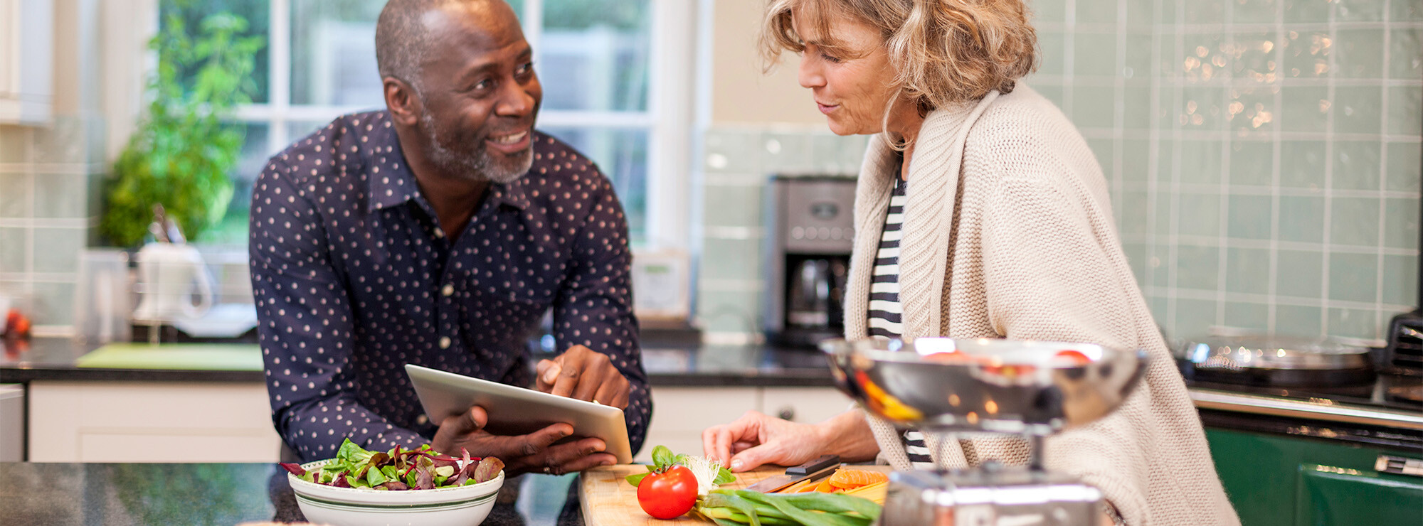 Un couple âgé consulte une tablette tout en préparant le dîner